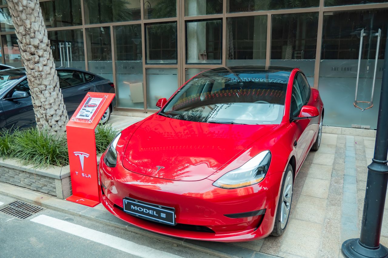 Shiny red Tesla Model 3 parked on sidewalk with reflections on windshield.
